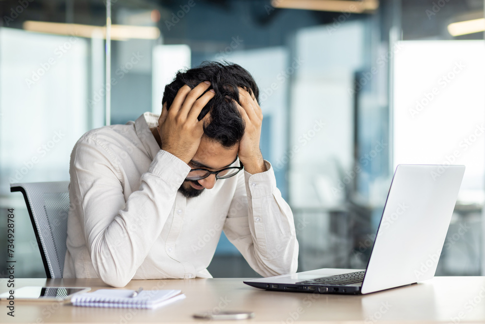 Frustrated and worried young Indian male businessman sitting in office at desk with laptop and hands on head, struggling to solve problem, tired Frustrated and worried young Indian male businessman sitting in office at desk with laptop and hands on head, struggling to solve problem, tired