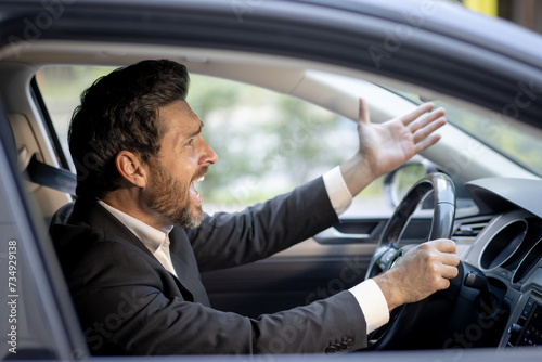 Close-up photo of angry young businessman in suit driving car and emotionally gesturing with hands, displeased shouting and arguing © Tetiana