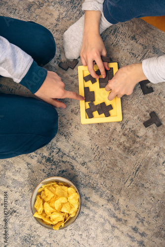 Top view of two people playing a wooden puzzle game on the floor with a bowl of chips nearby, enjoying a family weekend at home