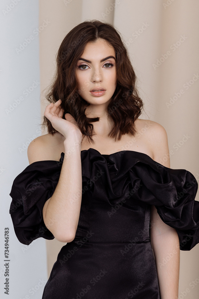Caucasian woman model in a stylish black dress with open shoulders against a background of cream curtains. Concept fashion show of dresses, designer clothing or shampoo and hair care products.