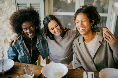 Cheerful woman sitting with arm around female friends at dining table enjoying dinner in patio photo