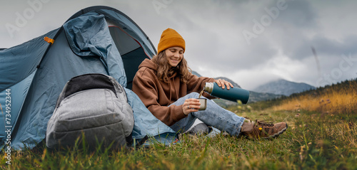 A young cheerful woman traveling with a tent pouring hot tea from a thermos. Hiking and trekking concept © olezzo