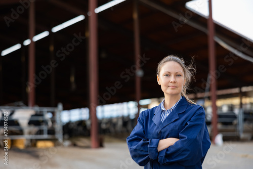 Portrait of woman ranch worker standing in cowshed near rows of cows in stalls with arms crossed. © JackF