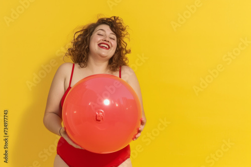 Funny young happy fat woman in red swimsuit having fun holding inflatable ball and going on summer holiday trip standing on yellow studio background. Vacation tour and travel concept.
