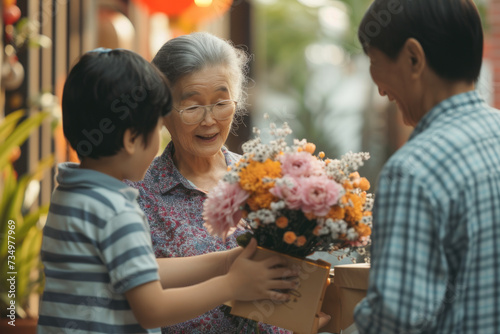 Happy Asian grandmother receives birthday presents from her loving family. Children together with grandfather give grandma a card and a bouquet of beautiful flowers © AI_images