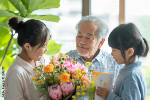 Happy Japanese grandfather receives birthday presents from his loving family. Children together with grandmother give grandpa a card and a bouquet of beautiful flowers