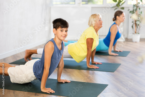 Diligent preteen boy practicing upward facing dog pose of yoga in fitness room with other persons