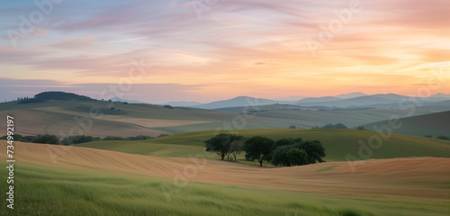 A serene countryside awakens to the soft hues of sunrise, painting the rolling hills and fields with a palette of pastel colors under a sky streaked with wispy clouds.