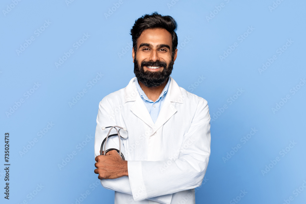 Smiling indian middle aged male doctor with crossed arms on blue backdrop Smiling indian middle aged male doctor with crossed arms on blue backdrop
