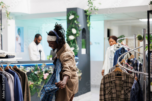 Cheerful african american man in headphones holding shirt on hanger while shopping in clothing store. Customer in good mood listening to music while choosing new outfit in fashion boutique