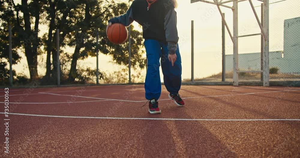 A blonde girl in a sports uniform maneuvers with the ball bouncing it off the floor during her game of basketball on the Red street court in the summer morning