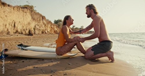Happy cheerful blonde girl in an orange swimsuit holds hands with her blonde boyfriend and sits on a surfboard on the sandy seashore with rocks in the morning at Sunrise