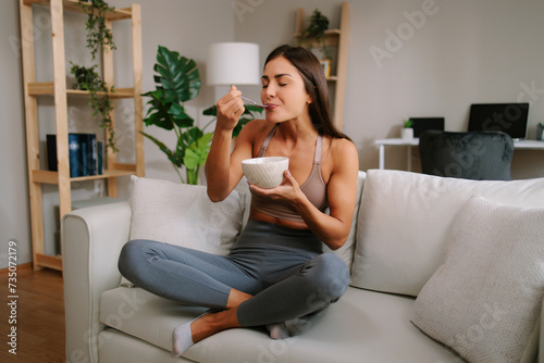 Beautiful young woman is sitting on the sofa in the living room and eating oatmeal