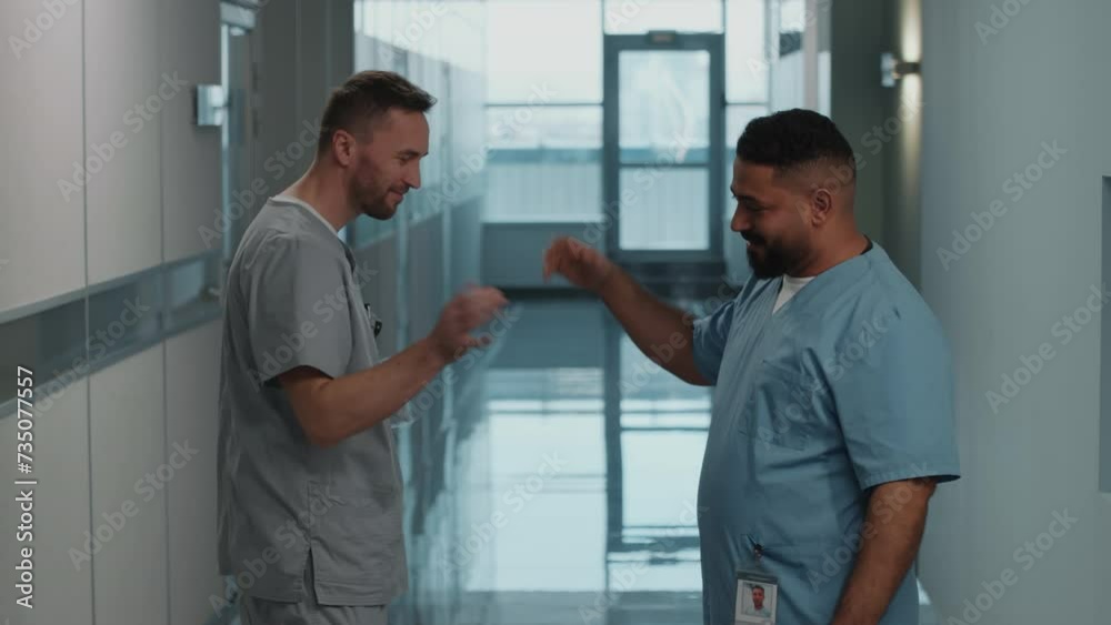 Two male hospital workers in scrubs greeting each other with fist bump, smiling and chatting while meeting in corridor during workday Two male hospital workers in scrubs greeting each other with fist bump, smiling and chatting while meeting in corridor during workday