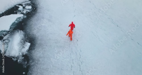 Two man in immersion suits walk Antarctica ocean ice. People explore South Pole. Group of scientists in orange wetsuits research climate and arctic wild nature. Aerial top trekking drone shot