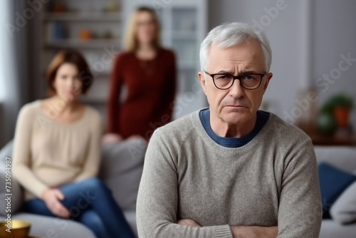 Senior caucasian spouses misunderstanding each other, experiencing crisis in relationship, offended wife looks disappointedly at camera, husband sits in defocus on background. Quarrel, marital crisis