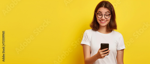 Mulher jovem surpresa em camiseta branca, segurando o smartphone e olhando para a câmera isolada em fundo amarelo photo
