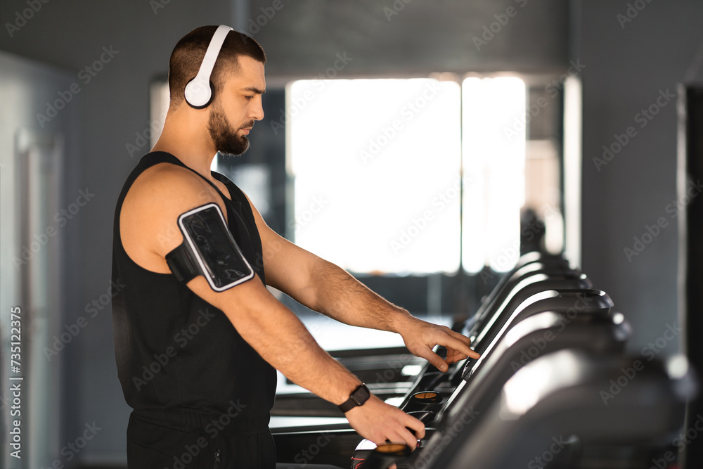 Focused young athlete with headphones and armband adjusting settings on treadmill Focused young athlete with headphones and armband adjusting settings on treadmill