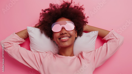 Retrato de uma linda mulher afro-americana com penteado afro, usando mascara para dormir cor de rosa e dormindo no travesseiro isolado sobre fundo rosa