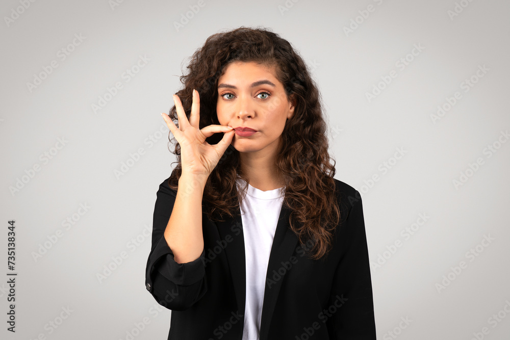 Young european businesswoman in black suit making silence gesture with her finger on her lips, grey backdrop Young european businesswoman in black suit making silence gesture with her finger on her lips, grey backdrop