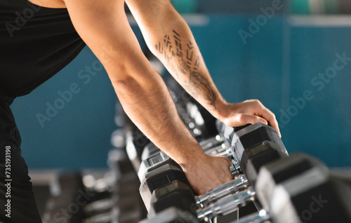 Sports Equipment. Unrecognizable man with tattoo taking dumbbells from gym rack