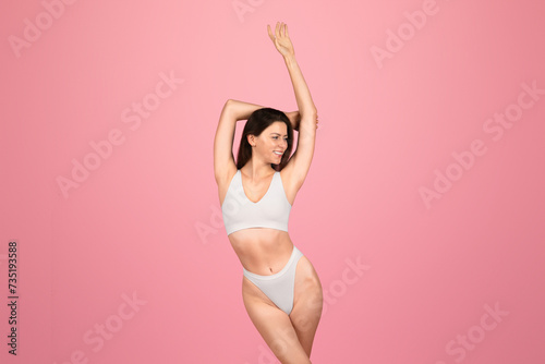 A cheerful woman with sleek brown hair enjoys a playful stretch in a minimalist white bikini © Prostock-studio