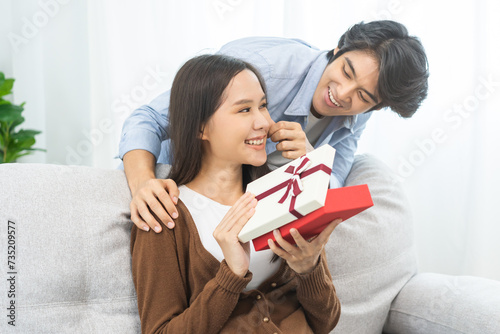 Celebrating Valentine's day anniversary, relationship two asian young couple love, boyfriend gives a gift to girlfriend by hide box at the behind, woman getting present while sitting on sofa at home.
