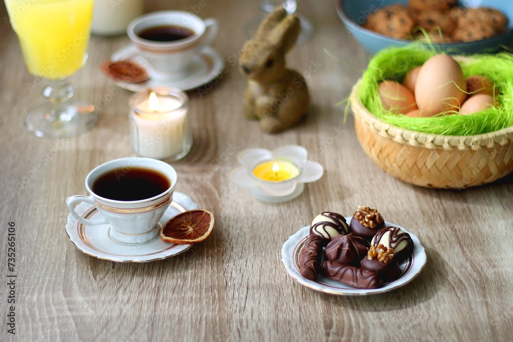 Easter eggs in the basket, bowl of cookies, chocolate pralines, Easter bunny figurine, cups of tea, glasses of juice, flowers and lit candles on the table. Selective focus. Easter eggs in the basket, bowl of cookies, chocolate pralines, Easter bunny figurine, cups of tea, glasses of juice, flowers and lit candles on the table. Selective focus.