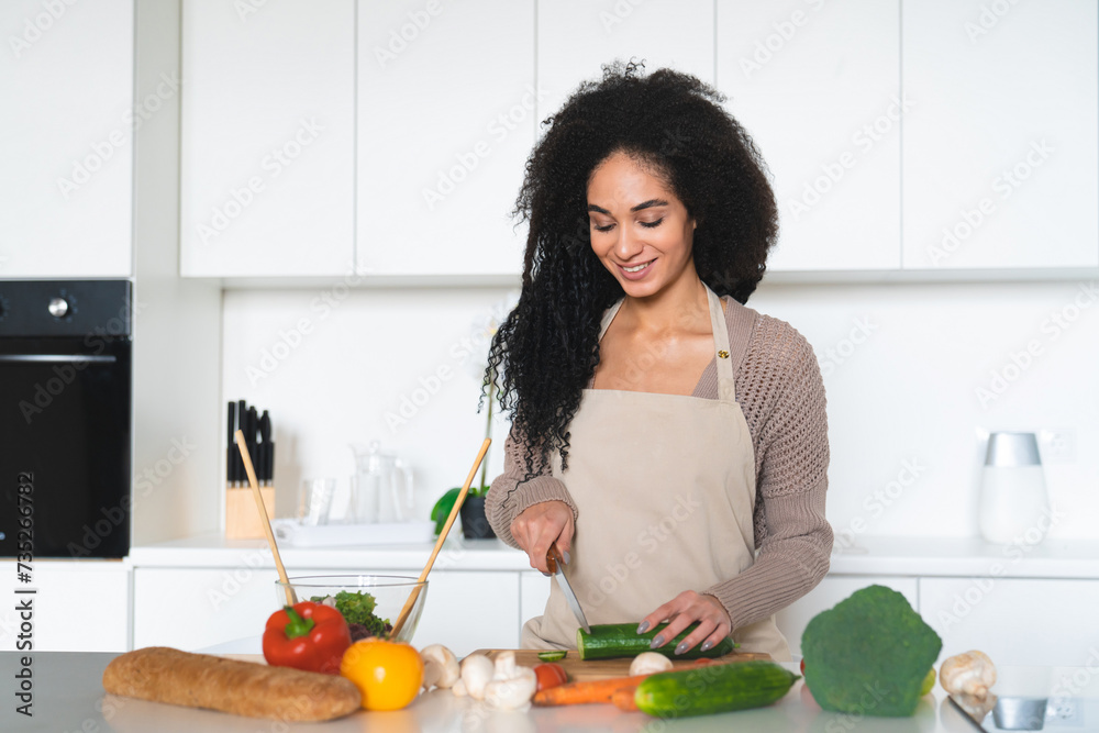 African young woman wearing apron, cooking salad, soup, vegetable vegan vegetarian dishes in the home kitchen, cutting cucumber. Healthy eating habits, dieting concept African young woman wearing apron, cooking salad, soup, vegetable vegan vegetarian dishes in the home kitchen, cutting cucumber. Healthy eating habits, dieting concept