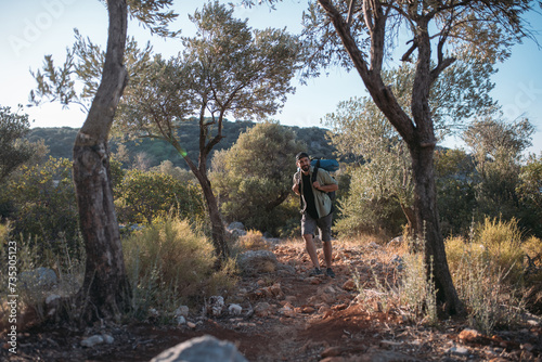 A male tourist with a large backpack and equipment on a mountain trail among the trees. © Anna