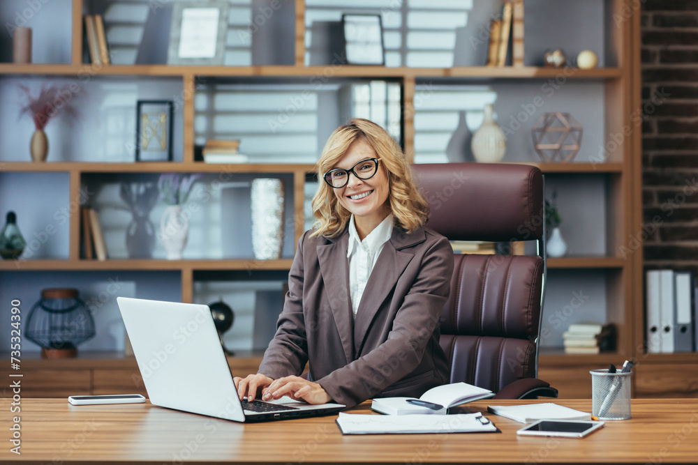 Portrait of a smiling senior business woman in a brown suit working in the office at a table on a laptop, looking and smiling at the camera. Portrait of a smiling senior business woman in a brown suit working in the office at a table on a laptop, looking and smiling at the camera.