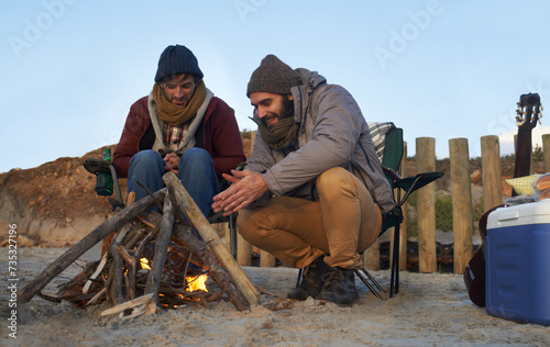Men, sunset and campfire with warming hands, smile and happy from travel adventure and journey outdoor. Friends, ocean and sea with bonding, hug and vacation by the beach on holiday with fire