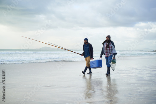 Winter, fishing and men walking on beach together with cooler, tackle box and holiday conversation. Ocean, fisherman or friends with rods, bait and tools in nature on morning vacation with cloudy sky photo