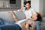 Man lying on couch with remote, woman sitting with cup, both smiling