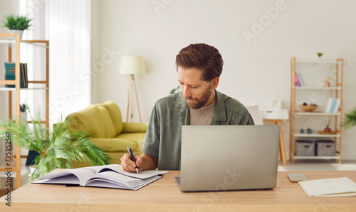 Portrait of a young business man in casual clothes working on a laptop computer at office or at home and writing at the desk making notes or calculations. Remote and freelance work concept. © Studio Romantic