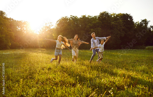 Happy family playing and having fun in nature. Cheerful, joyful mum, dad and children running together on a grassy lawn in a beautiful, fresh, green park on a warm, sunny summer evening