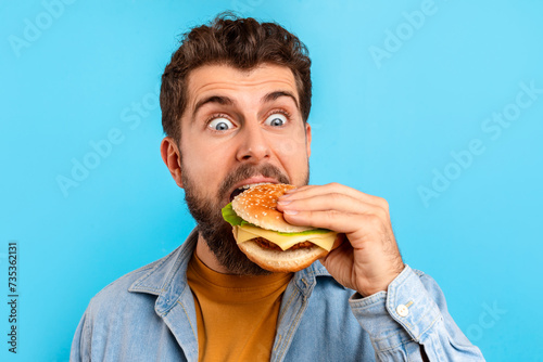 Hungry caucasian man on blue background excitedly biting into burger
