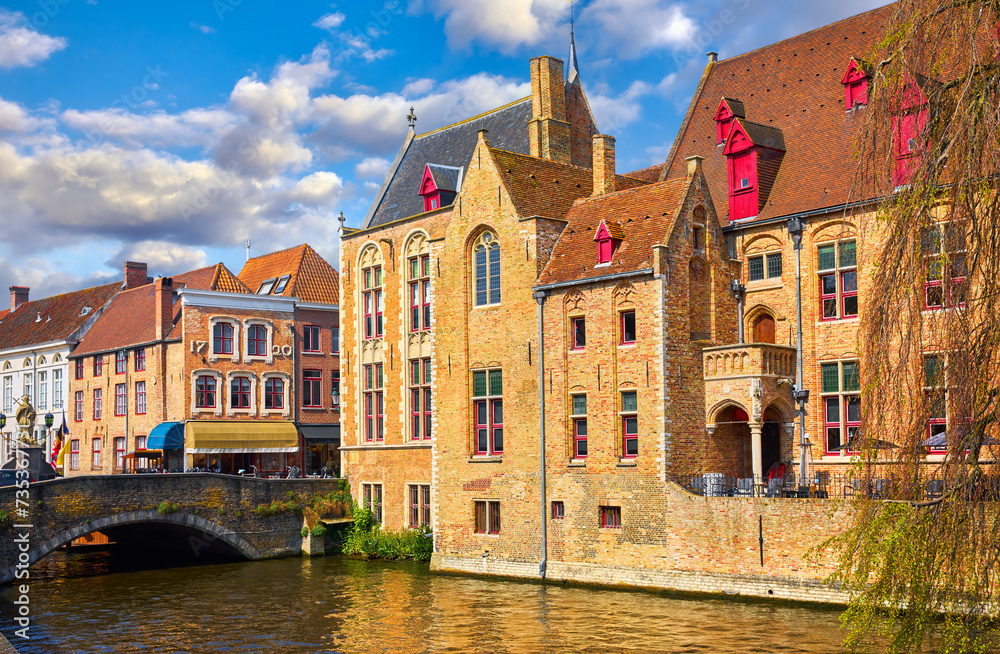 Bruges, Belgium. Ancient medieval european city. View at tower and vintage building on the banks of Rozenhoedkaai channel river. Old stone Bridge. Panoramic view with blue sky clouds. Famous tourist a