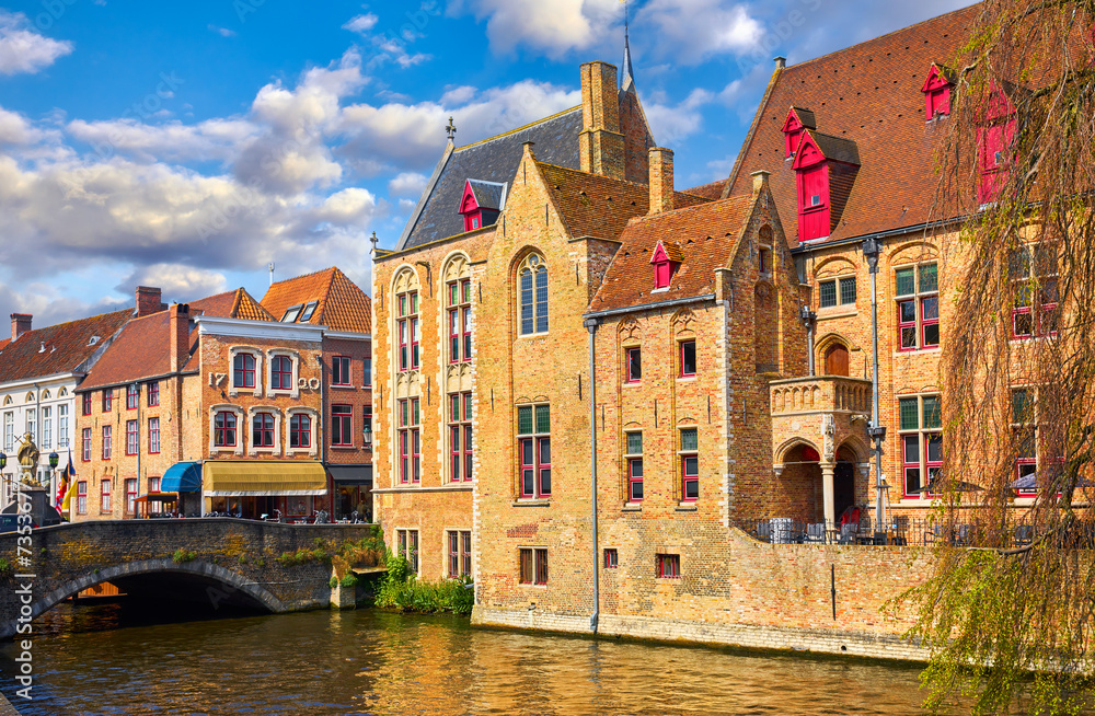 Fototapeta premium Bruges, Belgium. Ancient medieval european city. View at tower and vintage building on the banks of Rozenhoedkaai channel river. Old stone Bridge. Panoramic view with blue sky clouds. Famous tourist a
