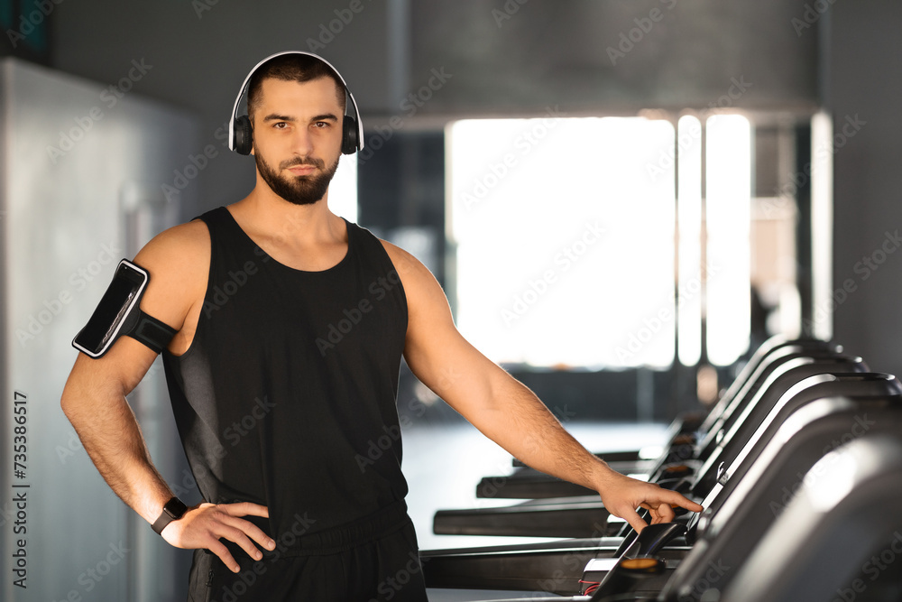 Confident young man with headphones and armband phone holder standing by treadmills Confident young man with headphones and armband phone holder standing by treadmills