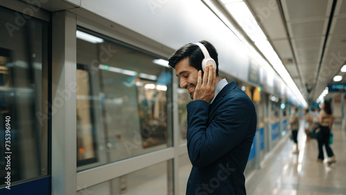 Project manager wearing headphone at train station while holding mobile phone for choosing song. Smart business man listening relaxing music while waiting for train with blurred background. Exultant.