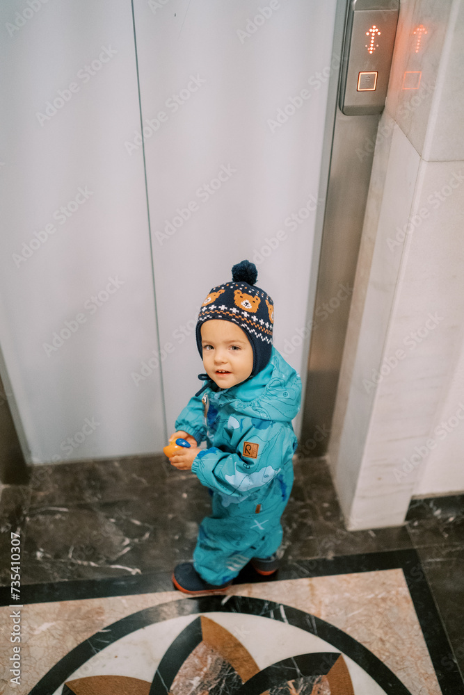 Little girl in overalls with a toy stands in front of the elevator door, looking back