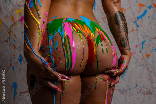 Attractive blonde female posing nude in front of paint splattered background with dripping paint in mulitple colors all over her body