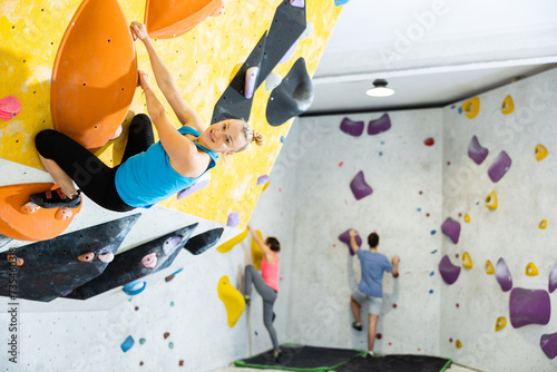 Woman looking at camera while climbing on rock-climbing wall during training in bouldering gym.