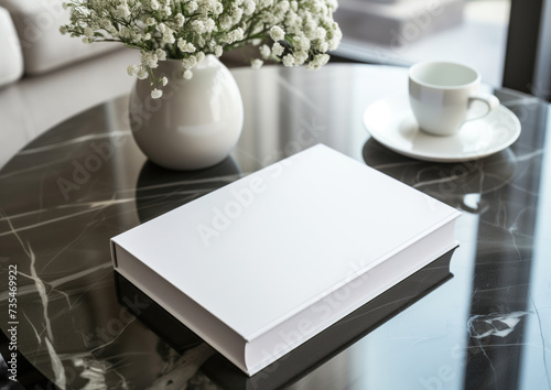 Advertising close up mockup shot of white book lying on black marble coffee table with coffee cup and flowerpot with flowers
