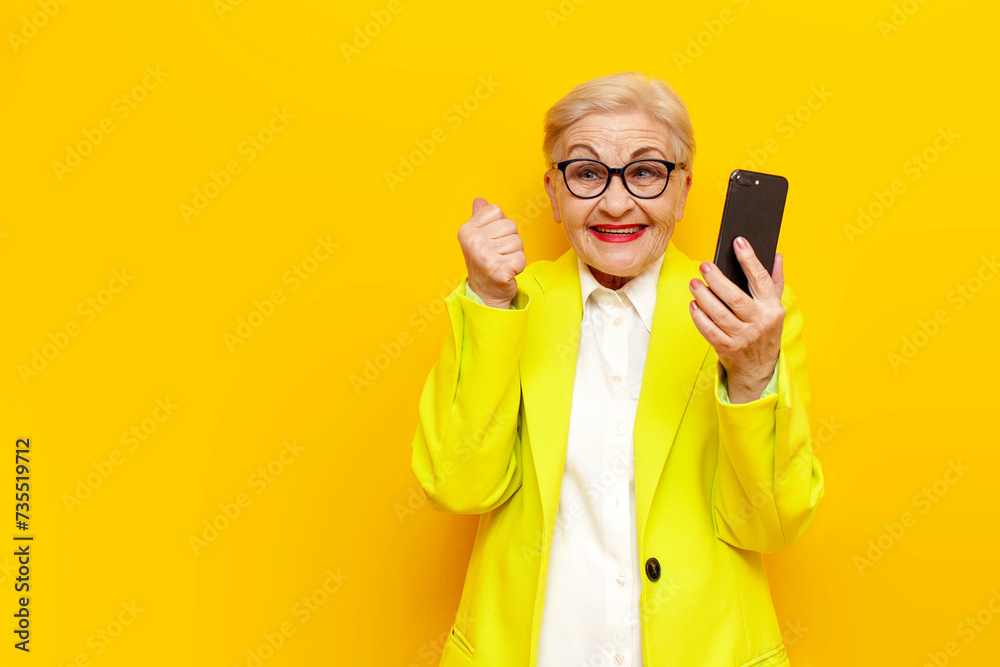 old businesswoman in glasses and formal wear uses smart phone and celebrates victory on yellow isolated background, elderly pensioner grandmother in blazer with mobile rejoices in success and luck old businesswoman in glasses and formal wear uses smart phone and celebrates victory on yellow isolated background, elderly pensioner grandmother in blazer with mobile rejoices in success and luck