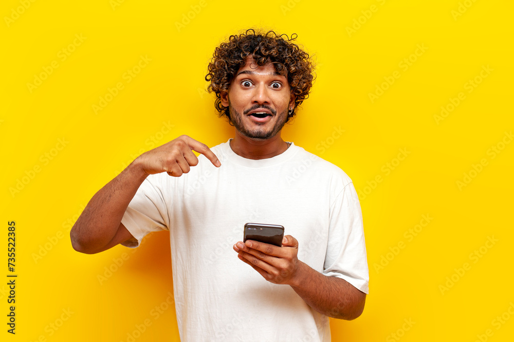 shocked curly indian man using smartphone online and pointing with hand on yellow isolated background, young surprised guy typing on phone in amazement shocked curly indian man using smartphone online and pointing with hand on yellow isolated background, young surprised guy typing on phone in amazement