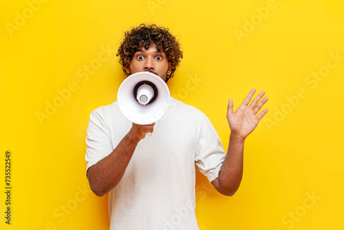 young Indian man in a white T-shirt announces information into a loudspeaker on a yellow isolated background, a curly-haired guy shouts into a megaphone