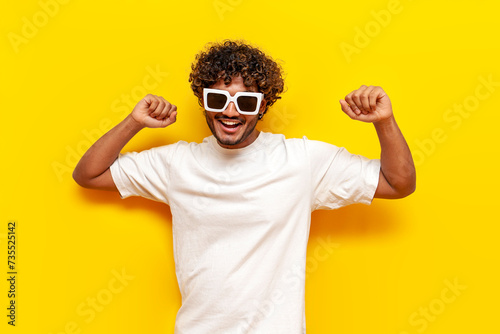 young cheerful indian man in white t-shirt and sunglasses dancing with raised hands on yellow isolated background  curly guy moving to music