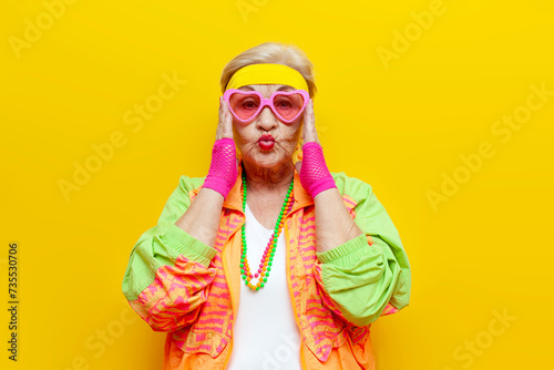 crazy funny old granny in sports colorful clothes and pink glasses on a yellow isolated background, elderly woman in youth hipster clothes looking at the camera